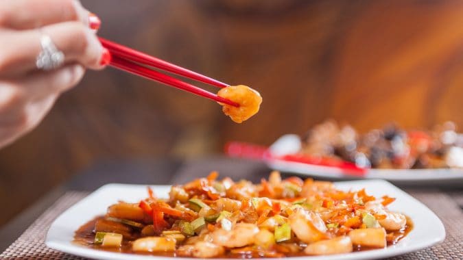 Mâncare chinezească cu legume și carne, servită cu bețișoare roșii, pe o farfurie albă.